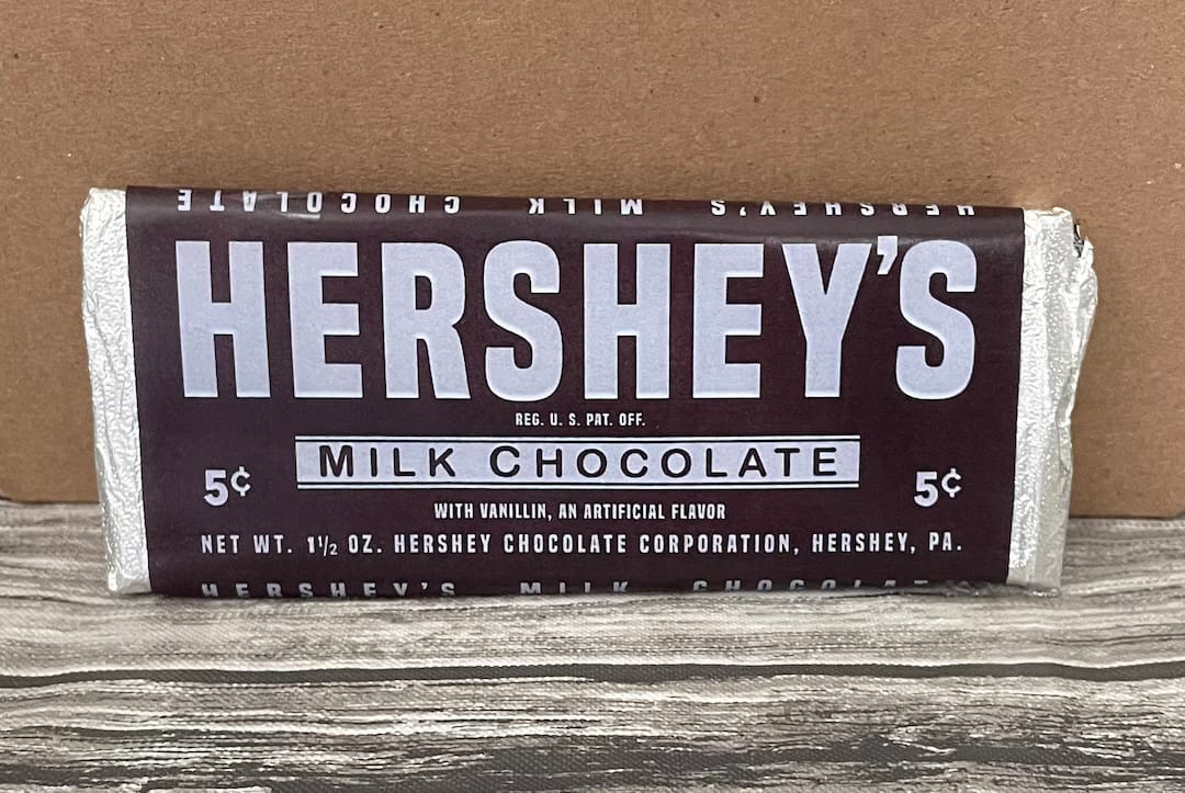 Photo of a Hershey's Milk Chocolate bar in a maroon and silver wrapper marked 5¢, propped against a cardboard backdrop.